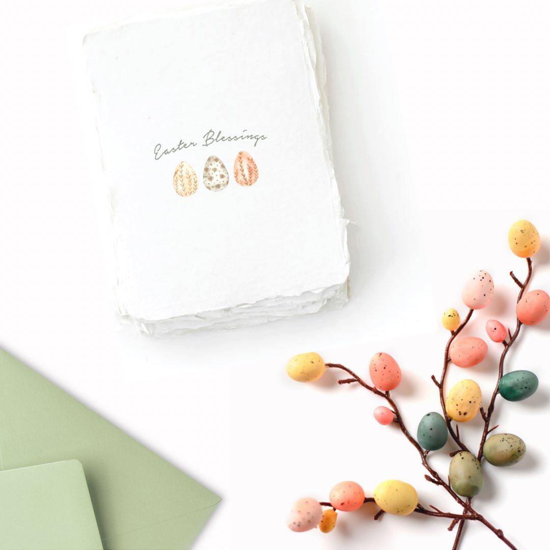 A flatlay image on a white background. In the top left, the focus of the image is a handmade card that reads Easter Blessings. There is a green envelope in the bottom left and colorful, spring easter eggs in the bottom right corner.
