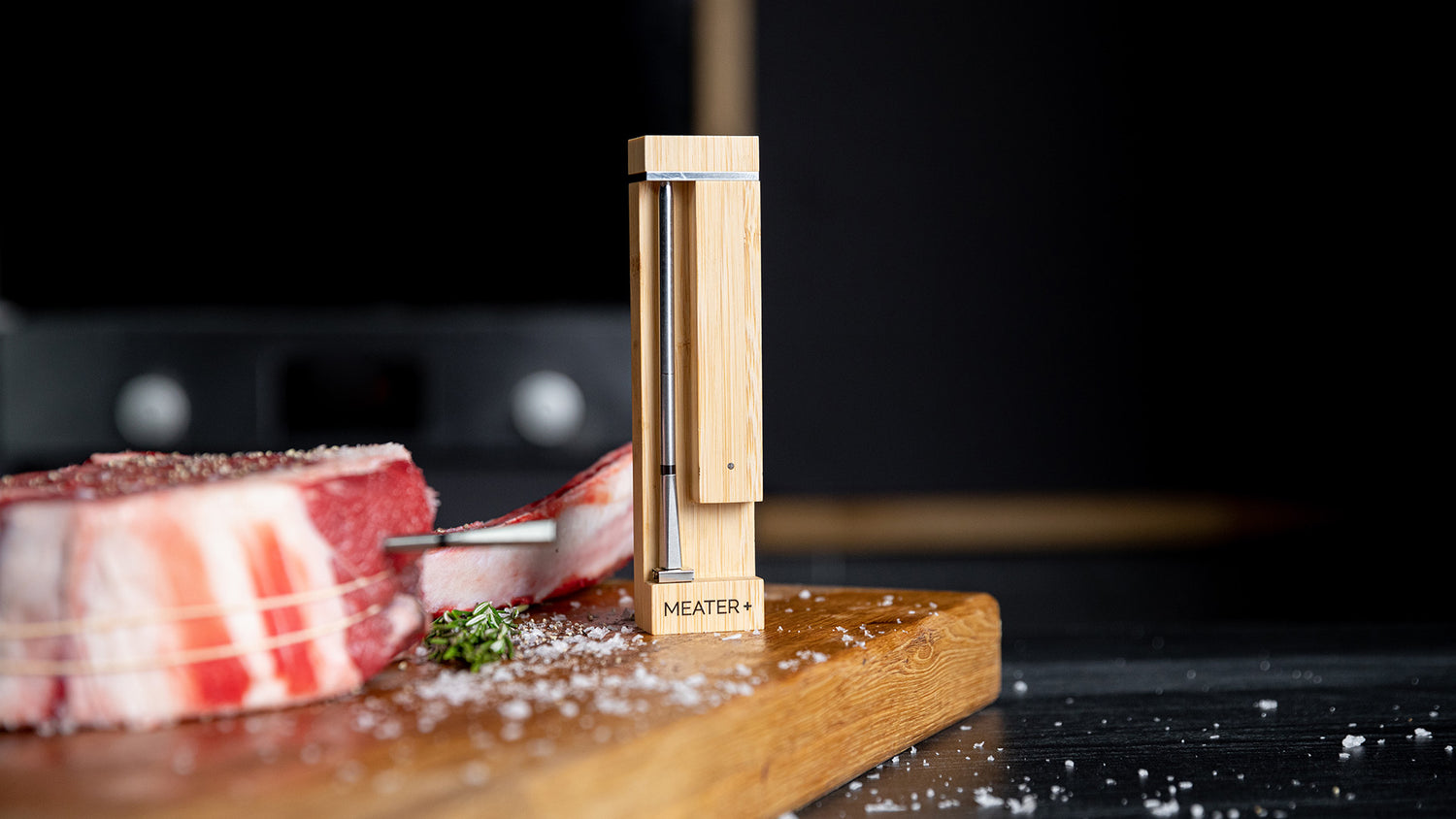 The new Meater 2 Plus thermometer in it's charging case sitting on a cutting board next to a large, uncooked steak.