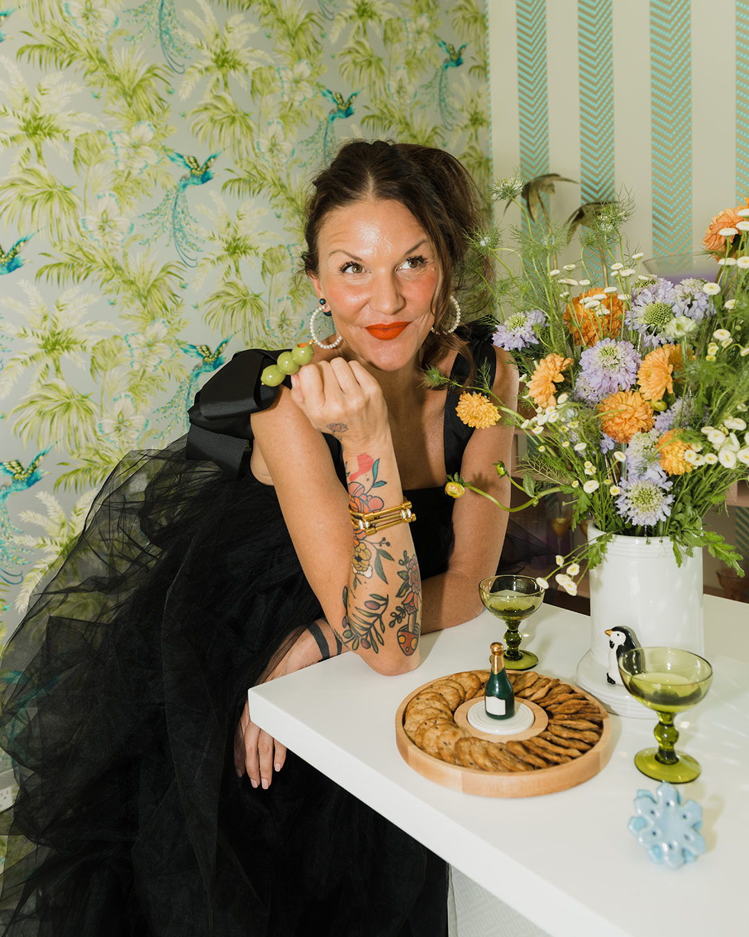 Woman sitting at a table with flowers and a cake, surrounded by decorative wallpaper.
